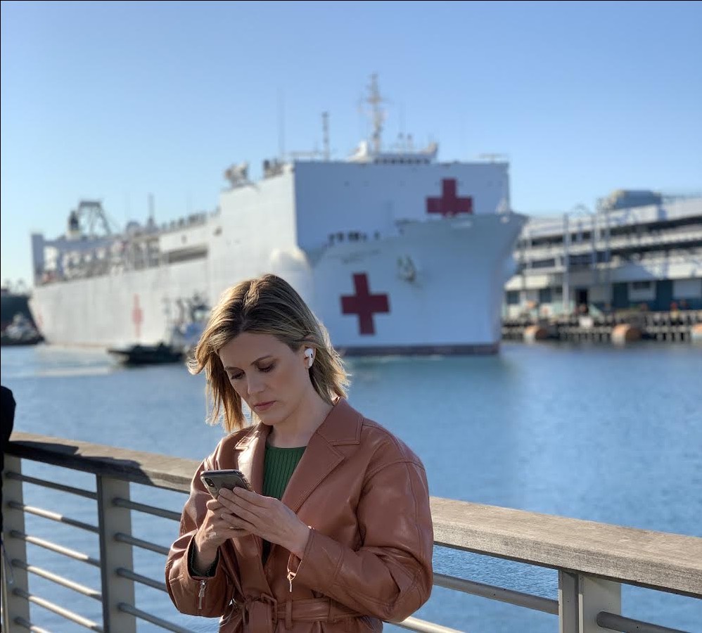 Erin McLaughlin checking her phone on a waterfront pier, a naval hospital ship moored behind her