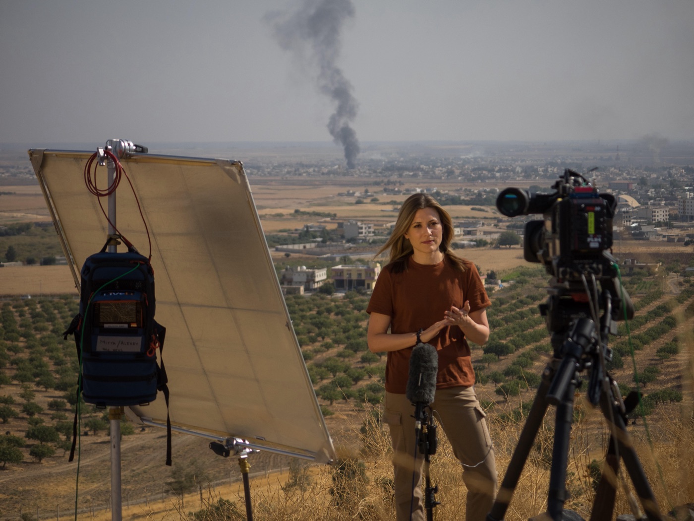 Erin McLaughlin at a live reporting position overlooking a town with smoke rising in the distance, beside a broadcast camera and lighting reflector
